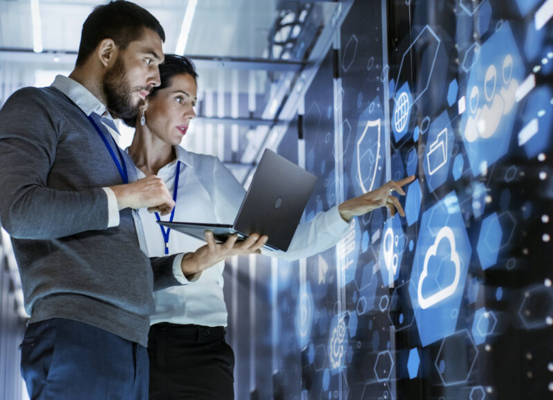 Male IT Specialist Holds Laptop and Discusses Work with Female Server Technician. They're Standing in Data Center, Rack Server Cabinet with Cloud Server Icon and Visualization.